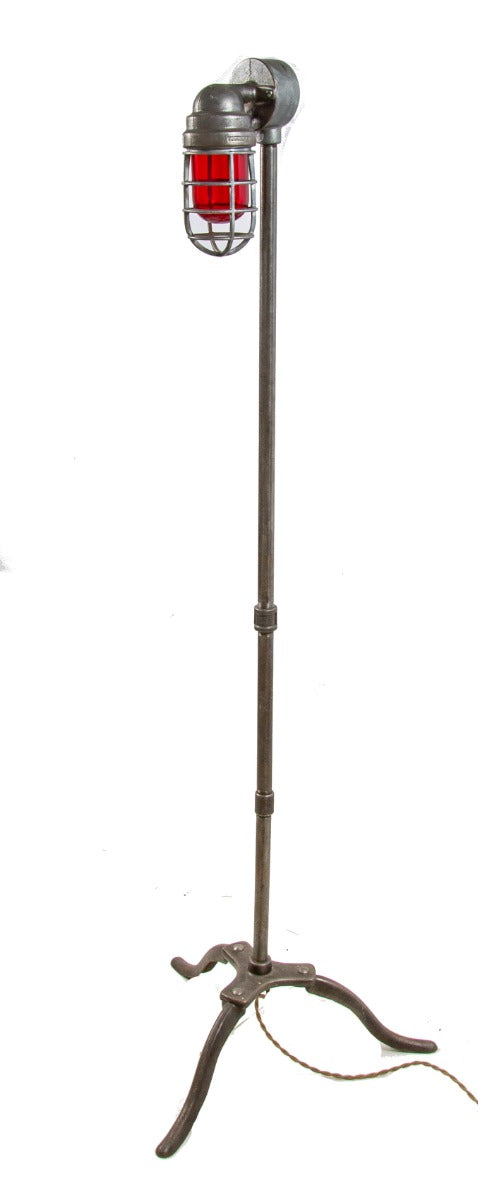repurposed brushed steel pipe floor lamp with three-legged cast iron base and ruby red explosion proof bulb guard